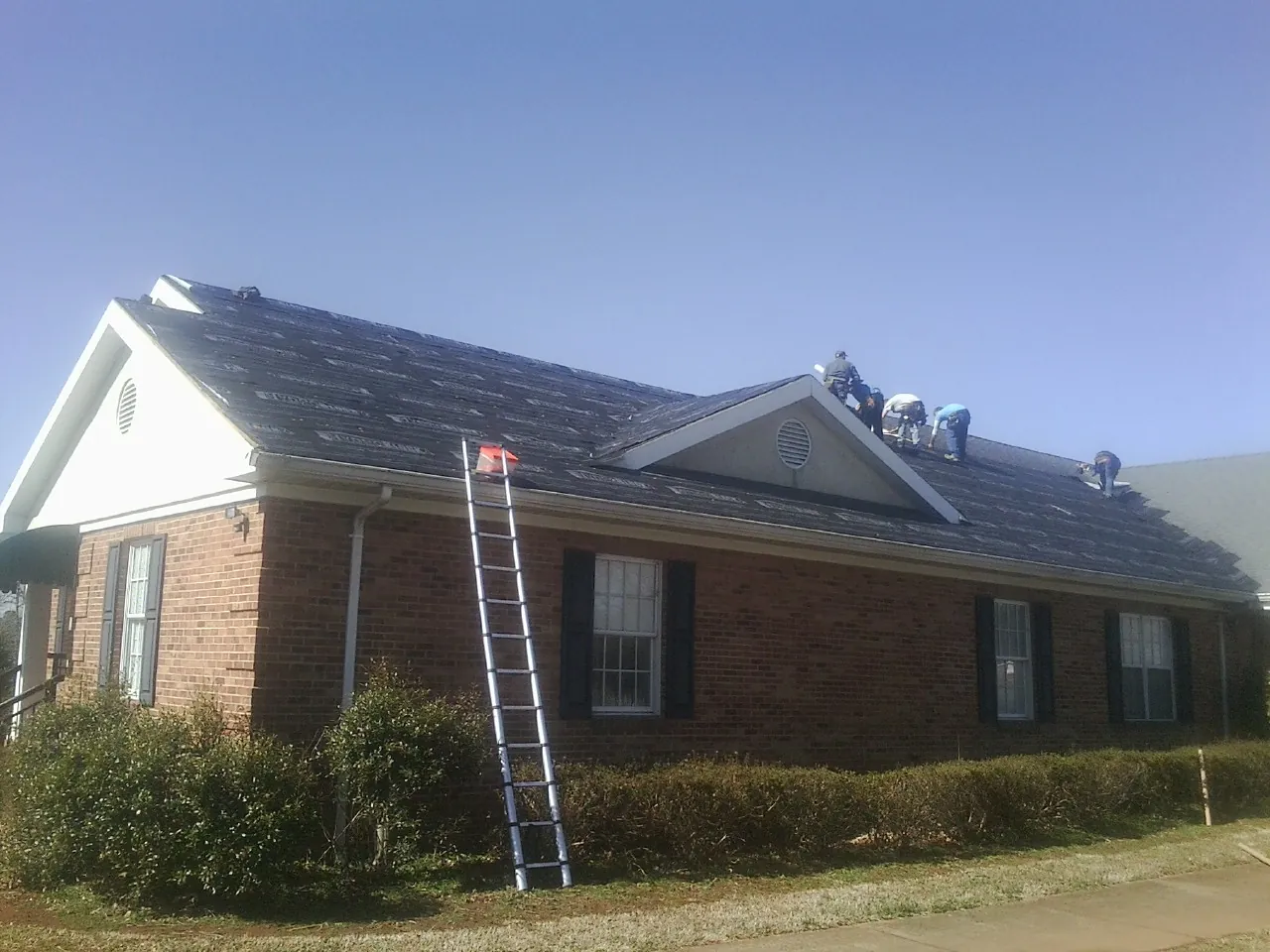 Crew tearing shingles off a brick ranch roof with a ladder set — Metal Roof Repair work by our crew in Colon