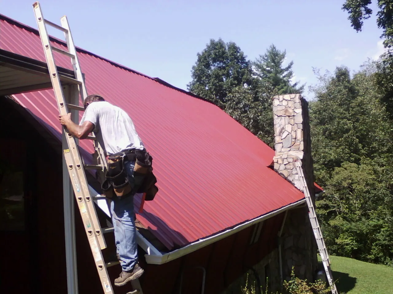 Roofer on a ladder beside a red metal roof and stone chimney — Soffit & Fascia Repair by Roofing Brothers in Colon